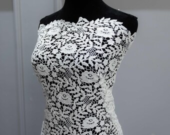 May include: A white lace fabric draped over a black mannequin. The lace features a floral pattern with intricate details. The fabric is likely intended for use in formal wear or bridal designs.