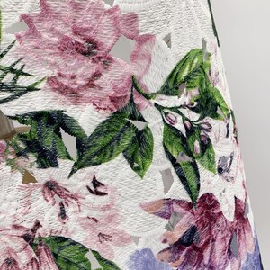 以下が含まれることがあります： 白い生地に花柄の刺繍が施されています。花はピンク、パープル、グリーンです。生地にはカットアウトパターンがあります。