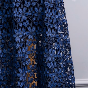 May include: A dark blue lace fabric featuring a repeating floral pattern with six-petaled flowers connected by intricate threadwork. The lace has a semi-sheer quality, revealing a solid dark base layer underneath the detailed embroidered floral design.