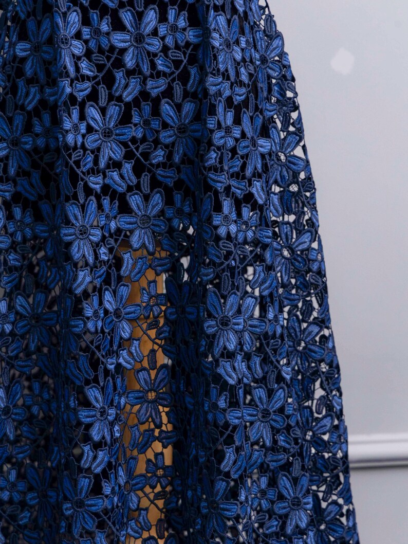 May include: A dark blue lace fabric featuring a repeating floral pattern with six-petaled flowers connected by intricate threadwork. The lace has a semi-sheer quality, revealing a solid dark base layer underneath the detailed embroidered floral design.
