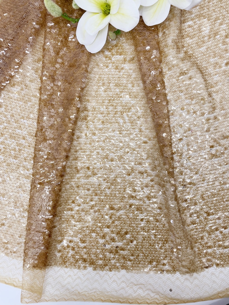 May include: A close-up of a gold sequined fabric with a delicate, sheer texture. The sequins are small and evenly spaced, creating a shimmering effect.