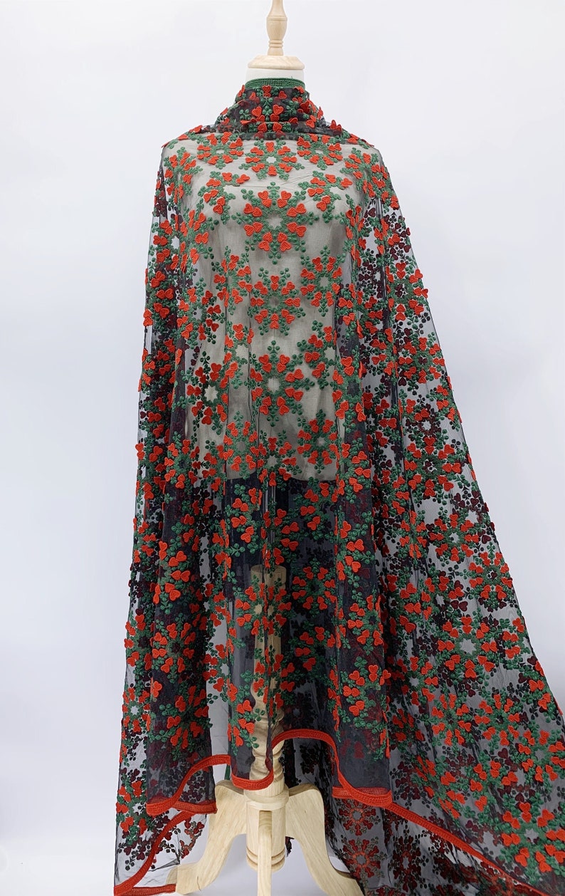 May include: Red and green floral embroidered fabric on a black mesh background. The fabric is draped over a mannequin and has a red trim around the edges.