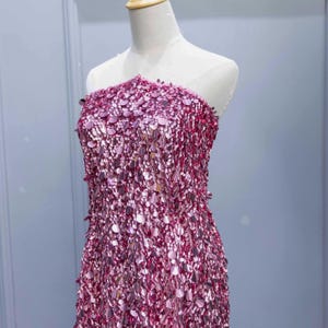 Könnte beinhalten: Ein paillettenbesetztes rosa Kleid auf einer Schaufensterpuppe. Das trägerlose Kleid reicht bis zu den Knien. Die Oberfläche ist mit kleinen, runden Pailletten bedeckt, die einen schimmernden Effekt erzeugen. Das Kleid ist für einen besonderen Anlass gedacht.