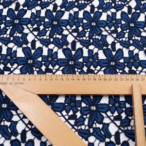 May include: A dark blue floral lace fabric featuring a repeating pattern of six-petaled flowers and leaves. A wooden ruler is placed across the fabric, showing measurements in centimeters up to 27 cm, with the text Made in China visible.