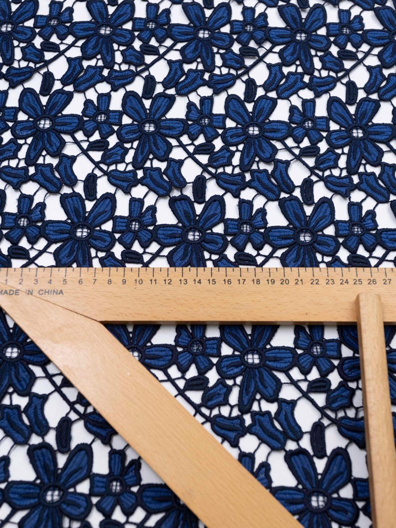 May include: A dark blue floral lace fabric featuring a repeating pattern of six-petaled flowers and leaves. A wooden ruler is placed across the fabric, showing measurements in centimeters up to 27 cm, with the text Made in China visible.