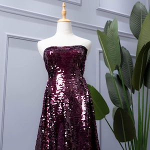 Può includere: Un abito lungo senza spalline su un manichino. L'abito è ricoperto di paillettes bordeaux scuro scintillanti. Le paillettes catturano la luce, creando un effetto scintillante. L'abito è esposto davanti a un muro grigio chiaro con una pianta sullo sfondo.