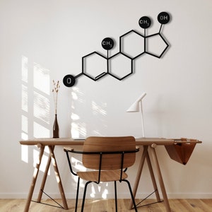 May include: A black metal wall decor in the shape of a chemical structure with the chemical formula CH3, CH3, O and OH. The decor is hanging on a white wall above a wooden desk with a chair.