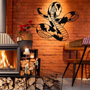 Op de afbeelding: Zwarte metalen wandkunst met een gekrulde slang met een gedetailleerd patroon. De kunst is gemonteerd op een bakstenen muur in een kamer met een open haard en een houten tafel met een stoel.