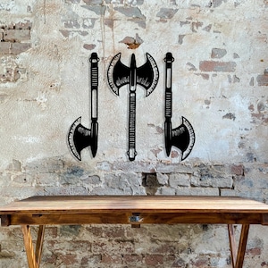 May include: Three black metal axe silhouettes mounted on a brick wall. The axes have a double-headed design with a pointed tip.