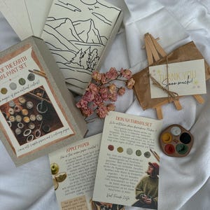 Könnte beinhalten: Ein Kunstzubehör-Set mit Naturfarben, Apfelpapier und Malvorlagen. Das Set enthält eine kleine Holzstaffelei mit einer Karte mit der Aufschrift "Thank You Sooo much!" und getrockneten Blumen.