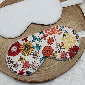 Op de afbeelding: Twee slaapmaskers. Eén is effen wit, de andere heeft een bloemenpatroon met rode, gele, oranje en blauwe bloemen op een crèmekleurige achtergrond. Beide maskers hebben elastische bandjes om ze om het hoofd te bevestigen.