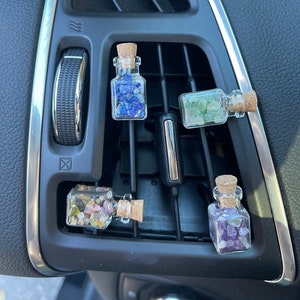 May include: Four small glass bottles with cork stoppers, each filled with colorful gemstones, are displayed in a car's air vent. The gemstones are blue, green, purple, and a mix of colors. The bottles are clear, allowing the gemstones to be visible.