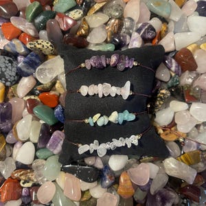 Puede incluir: Cuatro pulseras hechas con piedras preciosas de diferentes colores sobre un cojín de terciopelo negro. Las pulseras están hechas con amatista, cuarzo rosa, aguamarina y cuarzo transparente.