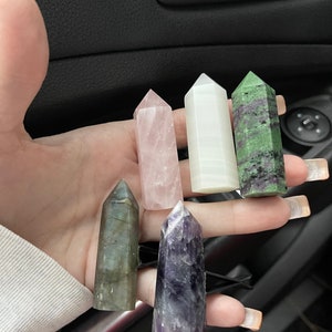 May include: Six different crystal points in various colors, including pink, white, green, gray, and purple. The crystals are arranged in a hand, with the points facing upwards.