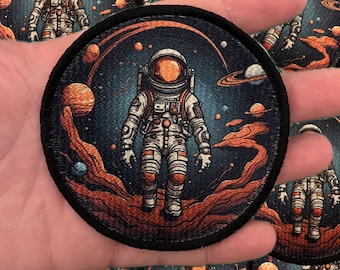 May include: A round embroidered patch featuring an astronaut in a white spacesuit standing on a rocky surface with a starry background. The astronaut is surrounded by planets and stars, and the patch has a black border.