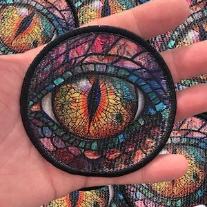 May include: A close-up of a colorful, embroidered patch featuring a large, detailed eye with a stained glass design. The eye is surrounded by a swirling pattern of colors, including shades of blue, green, orange, and purple.