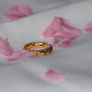 May include: A gold-colored ring with a textured design, resting on a white fabric surface. Pink flower petals are scattered around the ring, adding a touch of color and softness to the composition. The ring's intricate details are highlighted.