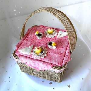 May include: A miniature wicker picnic basket with a pink floral tablecloth and a handle. The basket is open and the tablecloth is laid out with miniature teacups, saucers, and cupcakes.