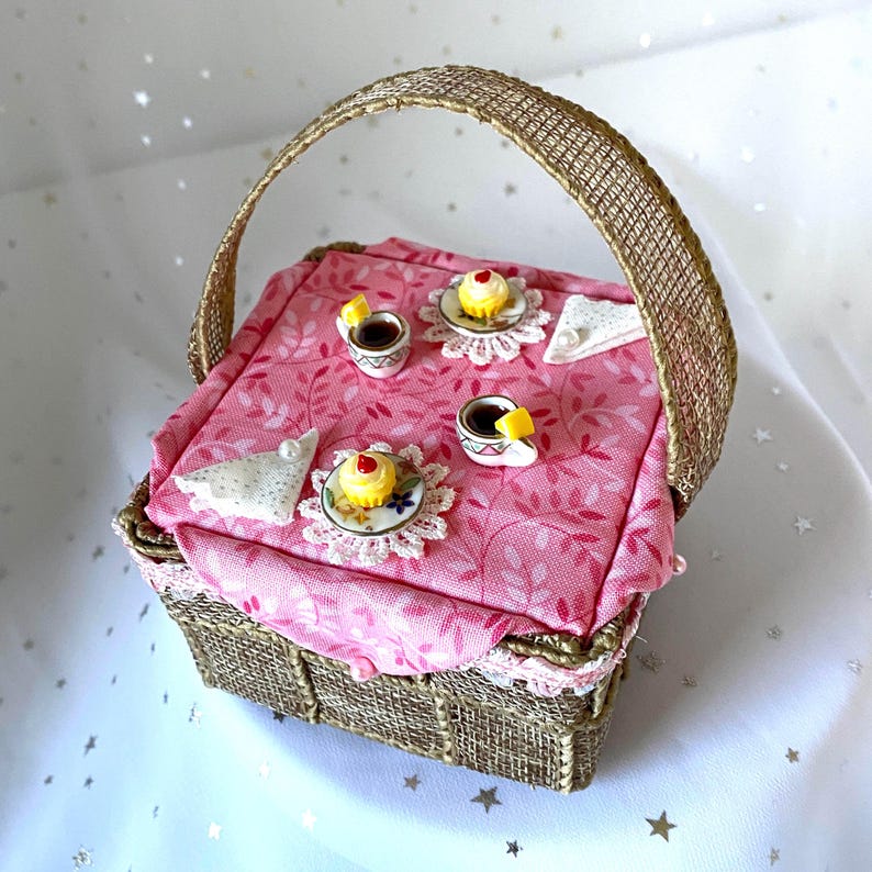 May include: A miniature wicker picnic basket with a pink floral tablecloth and a handle. The basket is open and the tablecloth is laid out with miniature teacups, saucers, and cupcakes.