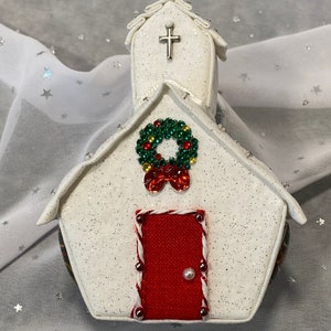 Pu&ograve; includere: Addobbo natalizio bianco a forma di chiesa con una porta rossa, una ghirlanda verde con accenti rossi e gialli e una croce argentata sul tetto.