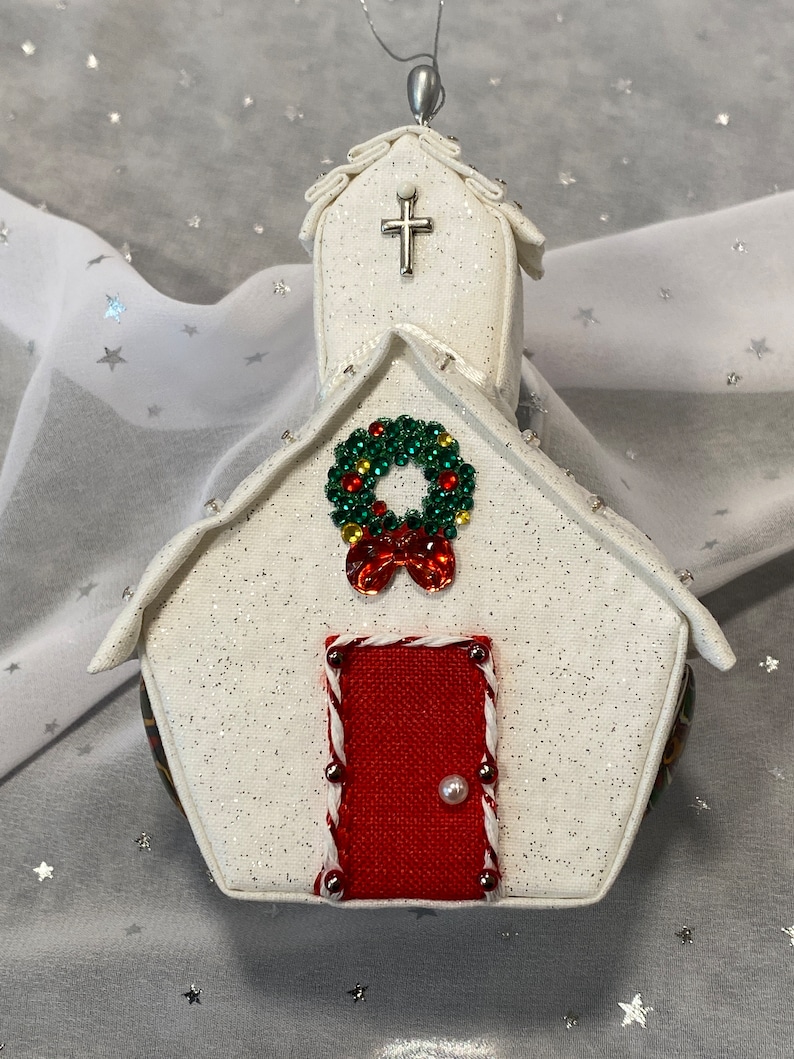 Pu&ograve; includere: Addobbo natalizio bianco a forma di chiesa con una porta rossa, una ghirlanda verde con accenti rossi e gialli e una croce argentata sul tetto.