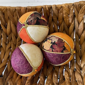 May include: Three decorative fabric balls with autumn leaf patterns, orange, purple, and cream sections, and gold trim. Each ball has a small decorative gold embellishment. The balls are displayed in a woven basket.