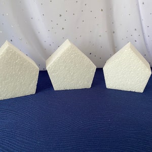 May include: Three white foam cake dummy shapes in the form of a house. The shapes are on a blue surface.