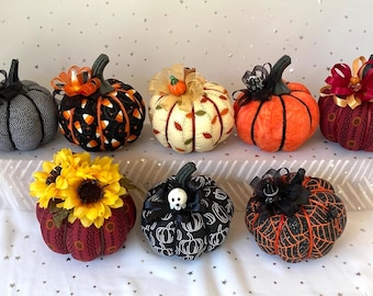 May include: A collection of decorative fabric pumpkins in various colors and patterns. Some pumpkins feature black and white polka dots, candy corn designs, and spiderwebs. Others are adorned with sunflowers, bows, and skull accents, perfect for fall or Halloween decor.