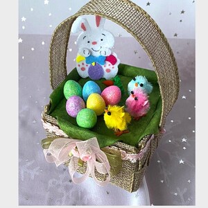 May include: A woven basket filled with Easter decorations. The basket contains colorful eggs, a white bunny, and small chicks. A pink ribbon with flowers adorns the front of the basket. The background is a light pink with white stars.