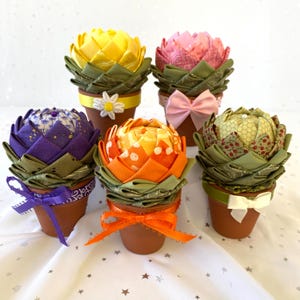 May include: Five fabric succulent planters in different colors, including yellow, pink, purple, orange, and green. Each planter is in a terracotta pot with a ribbon.