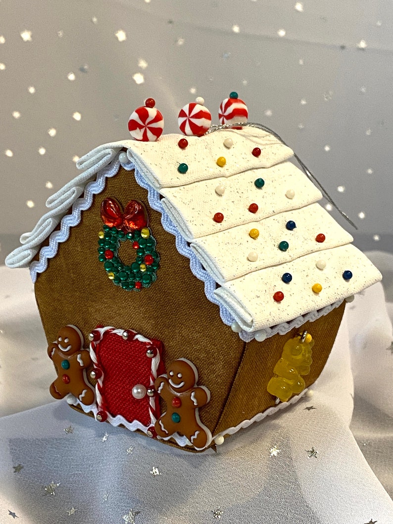 Pu&ograve; includere: Un ornamento per la casa di pan di zenzero marrone con glassa bianca, bastoncini di zucchero rossi e confettini colorati. La casa ha una ghirlanda sul davanti e omini di pan di zenzero sui lati.