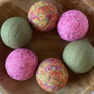 May include: Six fabric-covered balls in a wooden bowl. The balls are in various floral patterns, including pink, green, and orange.