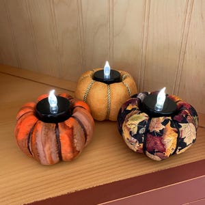 May include: Three decorative pumpkin-shaped candle holders with flickering LED lights. The pumpkins are in shades of orange, gold, and a fall leaf pattern. The candle holders are on a wooden shelf.