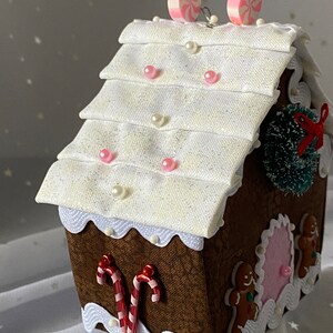 Pu&ograve; includere: Una decorazione per la casa di pan di zenzero con un tetto bianco, bastoncini di zucchero rosa e bianchi e personaggi di pan di zenzero. La casa &egrave; decorata con perline bianche e perline rosa.