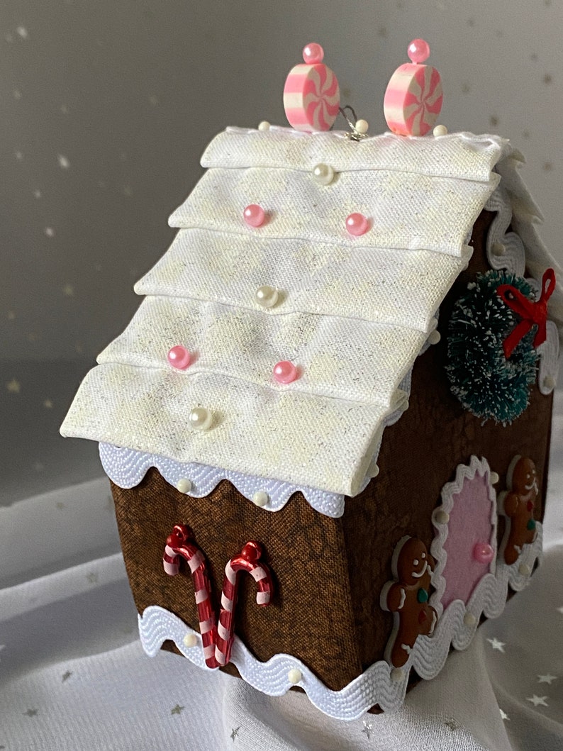 Pu&ograve; includere: Una decorazione per la casa di pan di zenzero con un tetto bianco, bastoncini di zucchero rosa e bianchi e personaggi di pan di zenzero. La casa &egrave; decorata con perline bianche e perline rosa.