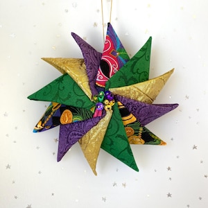 May include: A handmade star-shaped ornament with gold, green, and purple fabric. The ornament has a gold string for hanging and is decorated with small, colorful beads. The star's points are made of patterned fabric.