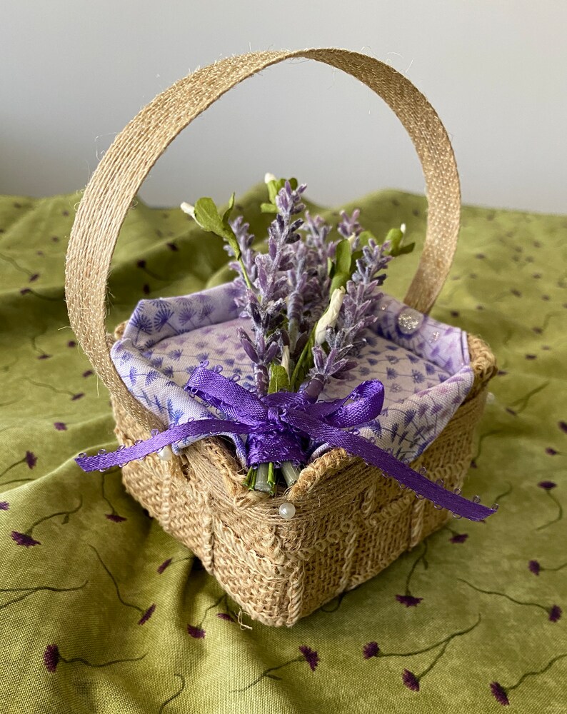 Pode incluir: Uma pequena cesta quadrada de juta com uma fita roxa e um buqu&ecirc; de flores de lavanda. A cesta tem uma al&ccedil;a de juta e &eacute; decorada com contas brancas.