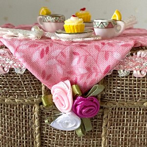 May include: A miniature tea party set with a pink and white checkered tablecloth, two teacups with lemon slices, a cupcake with pink frosting, and a floral arrangement with pink, white, and purple flowers.