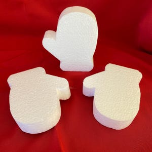 May include: Three white foam mitten shapes on a red background. One mitten is upright, and two are lying flat. The mittens are made of a textured foam material.