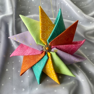 May include: A colorful fabric pinwheel ornament with a gold string for hanging. The pinwheel has eight points and is made of different fabrics in shades of green, yellow, orange, pink, and purple. A small gold and orange button is in the center of the pinwheel.