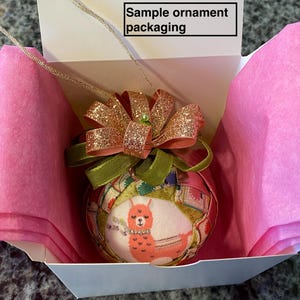May include: A pink and white gift box with a pink tissue paper lining. The box contains a round, fabric ornament with a llama design and a pink and gold bow.