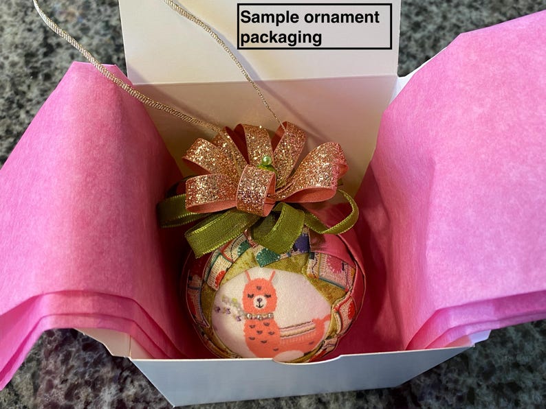 May include: A pink and white gift box with a pink tissue paper lining. The box contains a round, fabric ornament with a llama design and a pink and gold bow.