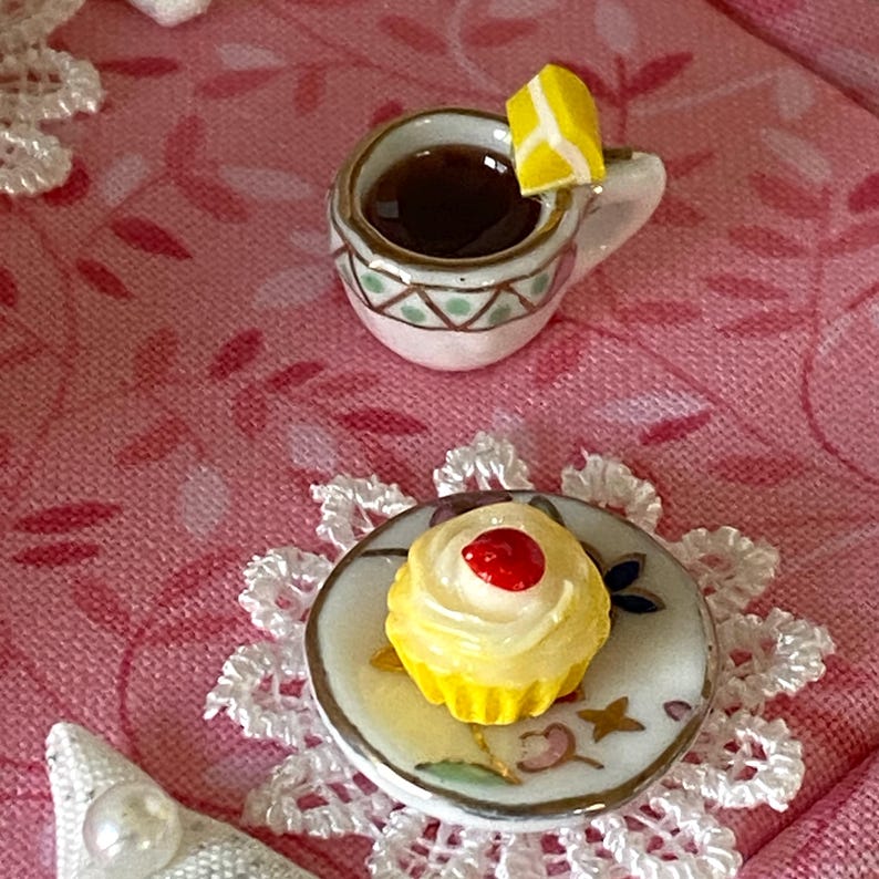 May include: A miniature teacup and saucer set with a yellow lemon wedge, a small cupcake with a red cherry on top, and a white lace doily on a pink background.