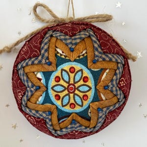 May include: A handcrafted Christmas ornament with a starburst design. The ornament features a burgundy background with a repeating pattern, and a central design in teal, gold, and blue. It is embellished with small blue beads and a jute twine loop for hanging.