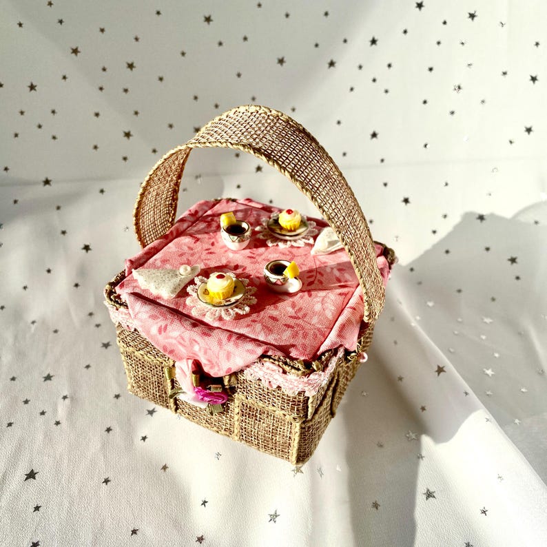 May include: A miniature picnic basket with a pink gingham tablecloth and a brown burlap handle. The basket is filled with miniature teacups and pastries.