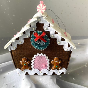 Pu&ograve; includere: Un ornamento per la casa di pan di zenzero marrone con glassa bianca, una canna di zucchero rosa in cima e due omini di pan di zenzero sul davanti. La casa ha una ghirlanda con un fiocco rosso e una porta rosa.