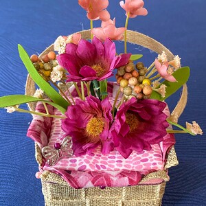 Pode incluir: Uma pequena cesta tecida com um forro de tecido estampado em rosa e branco. A cesta est&aacute; cheia de flores rosa e brancas e um pequeno amuleto de borboleta prateado.