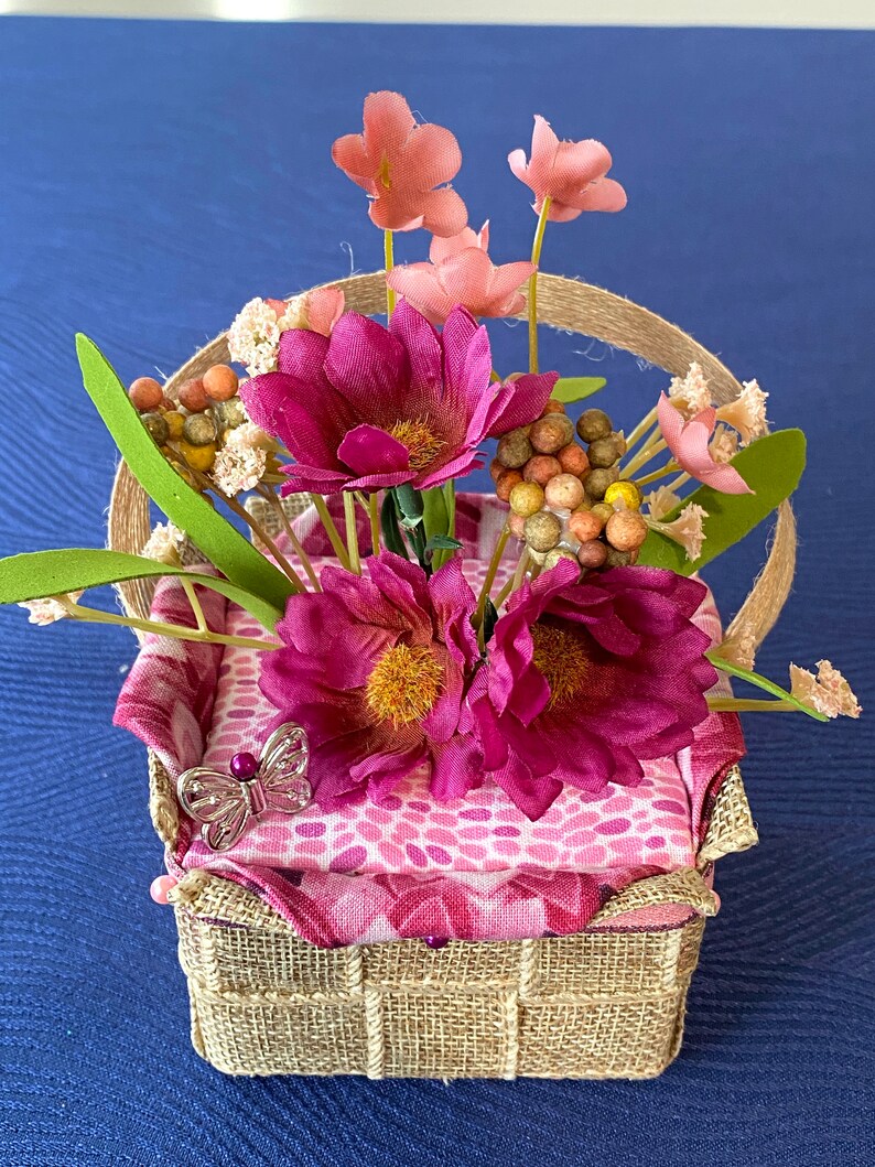 Pode incluir: Uma pequena cesta tecida com um forro de tecido estampado em rosa e branco. A cesta est&aacute; cheia de flores rosa e brancas e um pequeno amuleto de borboleta prateado.