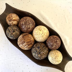 May include: A dark brown, elongated wooden bowl with curved ends containing eight decorative spheres. The spheres feature varied patterns including brown and tan wood grain, dark brown with gold circular motifs, and cream with leaf designs.