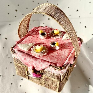 May include: A miniature picnic basket with a burlap handle and a pink fabric lining. The basket is filled with miniature teacups, saucers, and cupcakes.